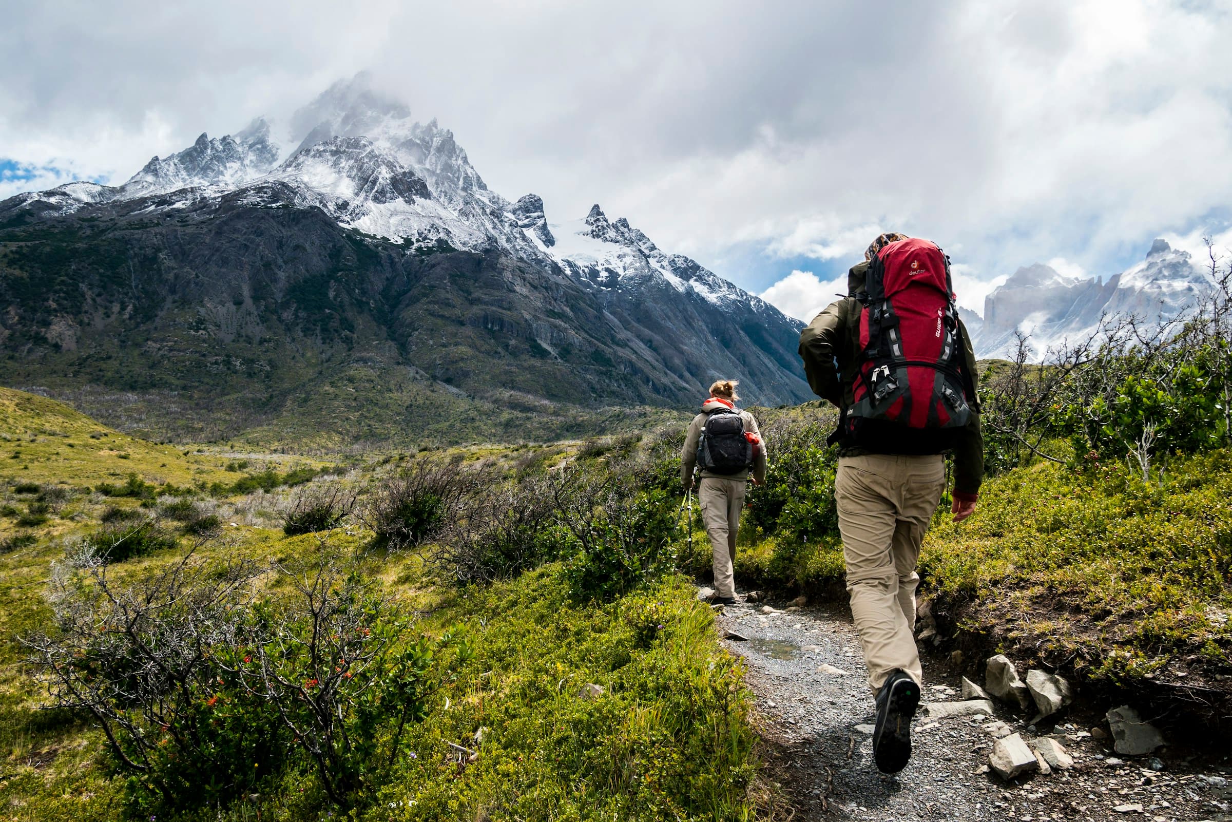 Two trekkers with packs walking a stone trail toward snow peaks